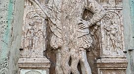 Epitaph des Hans Steininger in Braunau am Inn: Eine große Steinfigur in einer aufwendig verzierten Nische. Die Statue zeigt einen bärtigen Mann mit wehenden Haaren, der eine Robe trägt und eine Sichel in der Hand hält. Umgeben ist die Figur von komplexen Ornamenten und Reliefs mit Darstellungen von weiteren Figuren. Das Gesamtbild vermittelt einen feierlichen, historischen Eindruck.