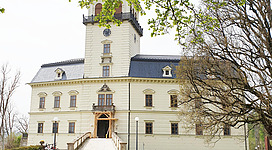 Ein historisches Gebäude mit einem hohen Turm und einer eleganten Fassade. Umgeben von Bäumen und einer ruhigen Atmosphäre.