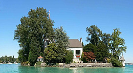 Schloss Litzlberg in Seewalchen am Attersee: Das Bild zeigt ein malerisches Haus, das sich auf einer Insel inmitten eines ruhigen Sees befindet. Das Gebäude ist von üppiger Vegetation umgeben und scheint ein traditionelles Landhaus-Styling zu haben. Im Hintergrund sind weitere Bäume und eine klare Himmelskulisse zu sehen.