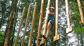 Ein Mädchen steht auf einem Kletterelement aus Holz zwischen Bäumen. Sie trägt einen Helm und einen Klettergurt.