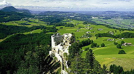Eine alte Burgruine auf einem Hügel, umgeben von grünen Wiesen und Wäldern. Im Hintergrund erstreckt sich eine weite, malerische Landschaft.