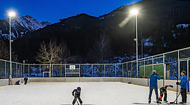 Ein Eishockeyfeld im Freien bei Dämmerung, umgeben von Bergen. Einige Kinder spielen auf dem Eis, während Erwachsene zuschauen.