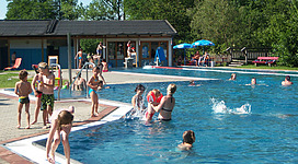 Freibad Maria Schmolln in Maria Schmolln: Eine Gruppe von Kindern und Erwachsenen genießt einen sonnigen Tag am Schwimmbad. Sie schwimmen, planschen und spielen im klaren Wasser des großen Beckens. Einige liegen auch auf Liegestühlen am Rand und beobachten die fröhlichen Aktivitäten. Das Schwimmbad ist von grünen Bäumen umgeben, die eine angenehme Atmosphäre schaffen.