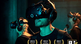 Eine Person mit einer Virtual-Reality-Brille und Kopfhörern erlebt ein immersives Abenteuer. Im Hintergrund sind weitere Teilnehmer zu sehen, die ebenfalls VR-Technologie nutzen.