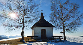 Eine kleine Kapelle steht auf einer schneebedeckten Wiese, umgeben von zwei Bäumen. Der Himmel ist klar und die Sonne scheint.
