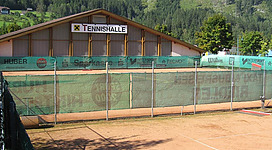 Eine Tennisanlage mit einem Sandplatz und einer großen Tennishalle im Hintergrund. Umgeben von grünen Bäumen und Bergen.