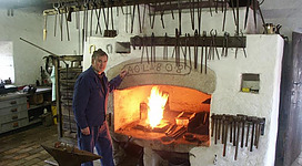 Hammerschmiede in St. Agatha: In einem gemütlichen Raum befindet sich ein großer Kamin, in dem ein loderndes Feuer brennt. An den Wänden hängen verschiedene Werkzeuge und Gerätschaften. Ein Mann, offenbar der Besitzer oder Betreiber des Raumes, steht neben dem Kamin und betrachtet das Feuer.