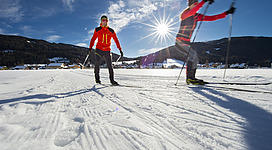 Zwei Skifahrer auf einer verschneiten Fläche unter einem klaren Himmel. Die Sonne scheint hell und erleuchtet die Szene.