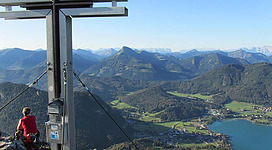 Ein Wanderer sitzt an einem Gipfelkreuz und blickt auf die umliegenden Berge und das Tal. Im Hintergrund sind grüne Wiesen und ein klarer See sichtbar.