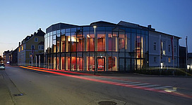 Ein modernes Gebäude mit großen Glasfenstern, das abends beleuchtet ist. Die Straße vor dem Gebäude zeigt Lichtspuren von vorbeifahrenden Autos.