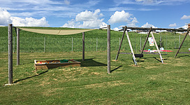 Freibad Kirchberg in Kirchberg ob der Donau: Eine Wiese mit Spielgeräten, eingerahmt von einem Zaun und umgeben von einem bewölkten Himmel. Auf der Wiese befinden sich Holzkisten und Klettergestelle, die für Aktivitäten oder ein Spielgelände zu dienen scheinen.