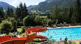 Ein Freibad mit einer großen Rutsche und vielen schwimmenden Menschen. Im Hintergrund sind grüne Berge und Bäume zu sehen.