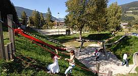 Kinder spielen bei Sonnenschein am Spielplatz, eine rote Rutsche sticht besonders hervor.