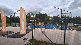 Padeltennis in Bad Ischl: Das Bild zeigt einen Outdoor-Sportplatz mit einem Netz-Tennisplatz in der Mitte. Der Bereich ist von Zäunen umgeben und mit Beleuchtung ausgestattet.