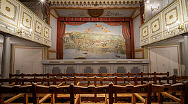 Das Historische Stadttheater Grein (1791) in Grein: Ein prunkvoll ausgestatteter Saal mit geschnitzter Holzvertäfelung, dekorativen Gemälden an den Wänden und einer üppigen, bemalten Decke. Die Anordnung von Holzbänken mit Nummern deutet auf einen Veranstaltungsort wie ein Theater oder eine Oper hin.