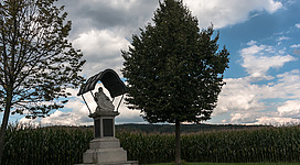 Das Steinerne Kreuz in Rufling in Leonding: Eine Statue eines Mannes in einer Nische, umgeben von Bäumen auf einer Wiese. Im Hintergrund ist ein bewölkter Himmel zu sehen.