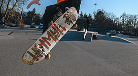 Skatepark Vöcklabruck - Cultural Boarders in Vöcklabruck: Ein junger Mann in oranger Kleidung steht auf einem Skateboard und führt eine Tricks auf einer schrägen Betonrampe durch. Er hebt das Skateboard hoch in die Luft und nimmt eine dynamische Körperhaltung ein. Im Hintergrund ist eine Parklandschaft mit Bäumen zu sehen.