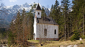 Eine kleine Kirche steht majestätisch inmitten eines Waldgebiets. Im Hintergrund sind schneebedeckte Berge und ein klarer Himmel zu sehen.