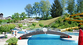 Erlebnisbad Mattighofen in Mattighofen: Eine Freizeitanlage mit einem großen Außenpool, der von grünen Rasenflächen, Büschen und Bäumen umgeben ist. Neben dem Pool gibt es mehrere Rutschen und andere Attraktionen, die die Anlage als Badepark oder Wasserpark erscheinen lassen. Das Gelände wirkt gut gepflegt und einladend.