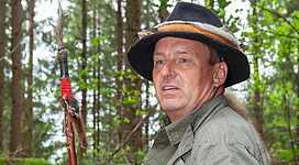 Waldbaden in Waldhausen im Strudengau: In diesem Bild sehen wir einen älteren Mann mit einem Hut, der in einer baumreichen Umgebung steht. Er hat einen ernsten Gesichtsausdruck und hält einen Stock in der Hand. Die Szenerie lässt auf eine ländliche oder waldreiche Gegend schließen.