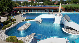 Erlebnisbad Eferding in Eferding: Eine große, offene Freischwimmbadanlage in einer idyllischen, grünen Umgebung. Mehrere Becken und Rutschbahnen mit klarem Wasser, umgeben von Terrassenstufen und Felsen. Ein ansprechendes Gebäude mit breiten Dachvorsprüngen und Liegen entlang der Poolränder vervollständigen das stimmungsvolle Bild eines modernen Badezentrums.