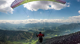 Ein Paraglider schwebt hoch über grünen Hügeln und Bergen. Der Himmel ist teils bewölkt mit vielversprechendem Wetter.