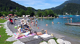 Strandbad St. Gilgen in St. Gilgen: Das Bild zeigt eine idyllische Szene an einem See. Viele Menschen genießen dort das sommerliche Wetter, indem sie im Wasser schwimmen oder auf dem Steg liegen und sich sonnen. Die Umgebung ist geprägt von grünen Wäldern und Bergen, die den See umgeben.