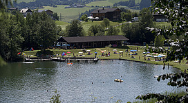 Ein schöner See umgeben von grünen Wiesen und Bäumen. Viele Menschen entspannen sich am Ufer und genießen die Sonne.