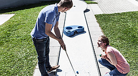 Ein Minigolfplatz im Freien mit zwei Personen. Die Frau beobachtet den Mann, während er seinen Schlag ausführt.