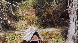 Eine kleine Kapelle aus Holz steht im Wald, umgeben von Bäumen. Neben der Kapelle befindet sich eine Bank zum Verweilen.