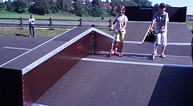 Skaterplatz hinter dem Viadukt in Seewalchen am Attersee: Das Bild zeigt einen öffentlichen Skatepark, der sich in einer grünen Umgebung mit Bäumen und Häusern befindet. Zwei Personen, vermutlich Jugendliche, fahren auf Rollern und Skateboards über die verschiedenen Rampen und Hindernisse.