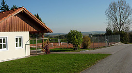 Tennisplatz Roßbach in Roßbach: Ein Landhaus mit roten Ziegeldächern und hellen Fassaden steht in einer grünen Umgebung. Vor dem Haus gibt es eine asphaltierte Straße und einen Tennisplatz mit Umzäunung. Im Hintergrund sind Bäume und eine hügelige Landschaft zu sehen.