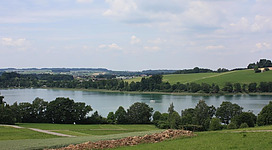 Fischen in Lochen am See in Lochen: Eine malerische Landschaft mit einem See, umgeben von grünen Hügeln, Wäldern und Wiesen. Ein Segelboot ist auf dem ruhigen Gewässer zu sehen, während eine Reihe von Gebäuden am Ufer und in der Ferne erkennbar sind. Die Szenerie strahlt eine friedliche und entspannende Atmosphäre aus.
