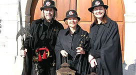 Eferding mit dem Nachtwächter in Eferding: In dem Bild sind drei Personen zu sehen, die traditionelle Kleidung mit Hüten tragen. Sie stehen vor einer großen Tür aus Holz mit einem gewölbten Eingang. Die Personen tragen schwarze Kleidung und halten in den Händen verschiedene Gegenstände wie eine Fackel und eine Laterne.