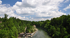 Traunfall in Roitham am Traunfall: Das Bild zeigt eine idyllische Landschaft mit einem Fluss, der sich durch dichten, grünen Wald schlängelt. Am Ufer sind einige Gebäude zu sehen, die sich harmonisch in die Umgebung einfügen. Der Himmel ist mit weißen Wolken bedeckt, die eine friedliche Stimmung erzeugen.
