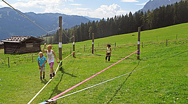 Kinder spielen auf einem großen grünen Wiesenfeld mit bunten Seilen. Im Hintergrund sind Berge und ein Holzschuppen zu sehen.