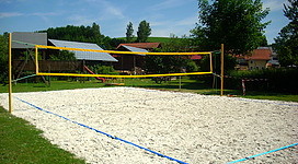 Beachvolleyballplatz in Oberkappel: Ein Volleyballfeld auf einer grünen Wiese. Es ist mit einem gelben und schwarzen Netz ausgestattet, das quer über das Feld gespannt ist. Rings um das Feld befinden sich einige Gebäude und Bäume. Der Himmel ist blau mit einigen weißen Wolken.