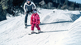 Ein Vater hilft seiner Tochter beim Skifahren auf einer verschneiten Piste. Die Tochter trägt eine rote Skijacke und einen Helm.
