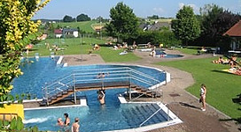 Freibad Frankenburg in Frankenburg am Hausruck: Ein Außenbereich mit einem Schwimmbecken, an dem mehrere Personen entspannen. Es scheint sich um einen Erholungsort zu handeln, der von Bäumen und Grünflächen umgeben ist.