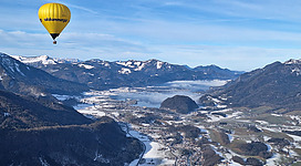 Der leuchtend gelbe Heißluftballon Salzkammergut schwebt hoch über einer malerischen Winterlandschaft. Der tiefblaue Himmel und die klare Sicht bieten einen atemberaubenden Blick auf die winterliche Idylle des Salzkammerguts.