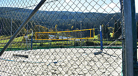 Ein Sandvolleyballfeld, umgeben von einem Zaun. Im Hintergrund sind Bäume und ein klarer Himmel zu sehen.