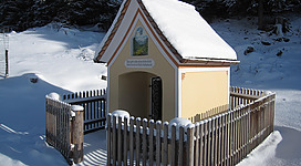 Eine kleine, hübsche Kapelle im Schnee, umgeben von einem Holzzaun. Die Dächer sind mit einer Schneedecke bedeckt und die Umgebung ist winterlich und ruhig.