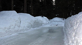 Eine gefrorene Straße umgeben von hohen Schneewänden. Die Umgebung ist bewaldet und vermittelt eine winterliche Atmosphäre.