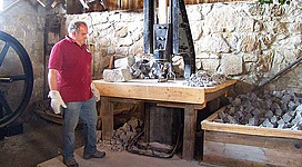 Granitlehrpfad Erlebniswelt Granit und Arena Granit in St. Martin im Mühlkreis: Eine Person in einem roten Hemd steht in einer Werkstatt mit einer Holzbank, auf der verschiedene Werkzeuge und Gesteinsbrocken liegen. Die Wände sind aus Stein, und an der Decke hängen weitere Werkzeuge. Rechts sind Regale aufgestellt, auf denen weitere Steine und Materialien gelagert sind.