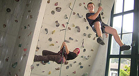 Zwei Kletterer an einer Kletterwand in einer Halle. Einer hängt in der Luft, während der andere sich am Seil abseilt.