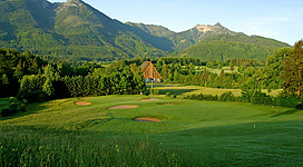 Golfclub Salzkammergut in St. Wolfgang im Salzkammergut: Das Bild zeigt eine malerische Landschaft mit einem prächtigen Berghang im Hintergrund. Im Vordergrund befindet sich eine große Rasenfläche mit Golfbunkern, die von dichtem Baumbestand umgeben ist. In der Mitte des Bildes ragt ein charakteristisches Gebäude mit traditioneller Architektur empor, das den Mittelpunkt der Szene bildet.