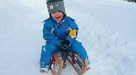 Ein kleines Kind sitzt auf einem Schlitten im Schnee. Es trägt eine blaue Winterjacke und einen grauen Hut und lächelt fröhlich.