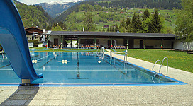 Ein Schwimmbad mit blauer Rutsche und klarem Wasser. Im Hintergrund sind grüne Hügel und Berge zu sehen.