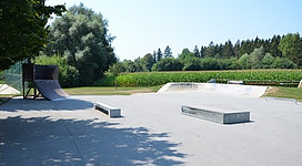 Funpark Schärding in Schärding: Ein Außensportbereich mit einer Rampe und einigen Betonblöcken, umgeben von Bäumen und Vegetation. Der Boden ist mit Beton oder Asphalt bedeckt und bietet möglicherweise Möglichkeiten für Aktivitäten wie Skateboarding oder andere Freizeitaktivitäten.