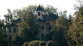 Schloss Auhof bei Perg in Perg: Ein malerisches Landhaus inmitten eines üppigen, grünen Gartens. Das zweistöckige Gebäude zeigt eine traditionelle Architektur mit einem spitzen Dach und dekorativen Elementen. Die Umgebung ist von Bäumen und Sträuchern umgeben, was dem Anwesen einen idyllischen Charakter verleiht.